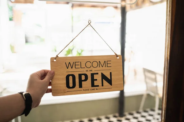 Hand woman hold open sign broad through the door glass. OPEN sign board through the glass of store window. text wooden board  door vintage shop sign opened store.