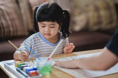 Küçük sevimli Asyalı bebek gülümsüyor ve annesi suluboya kullanarak renkli resimlerle resim yapıyor. Boyalı fırça rengini kullanan Asyalı bir kız. Bebek aktivitesi yaşam tarzı konsepti.