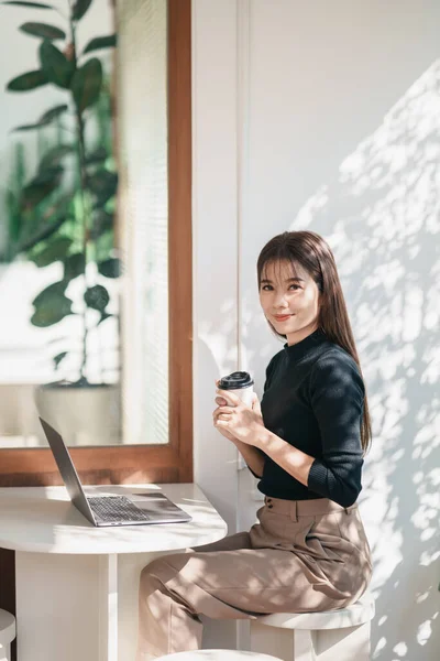 Asian freelance woman smiling holding cup of hot coffee and working on laptop computer on wooden table at cafe. Entrepreneur woman working for her business at coffee shop. Business work from anywhere