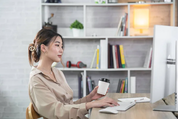 Asian freelance woman smiling holding cup of hot coffee and working on laptop computer on wooden table at home. Entrepreneur woman working for her business at home. Business work at home concept.
