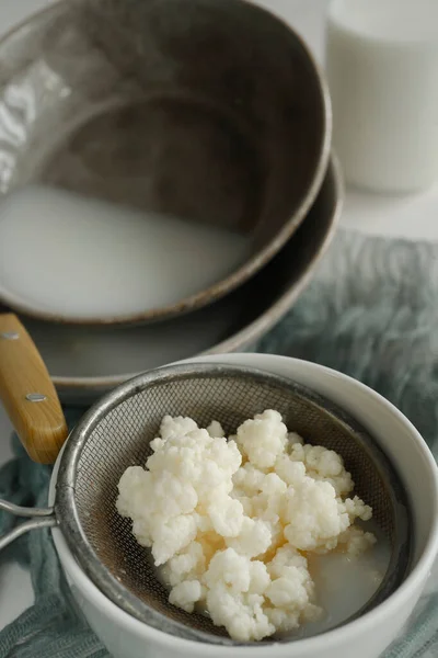 Grani di kefir di latte in un colino con piatti grigi su tavolo bianco. — Foto Stock