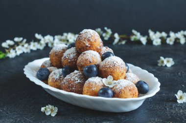 Ev yapımı süzme peynirli çörekler pudra şekeri ve kiraz çiçekli, gri bir arka planda böğürtlenli. Gün ışığı, tonlu resim, seçici odak
