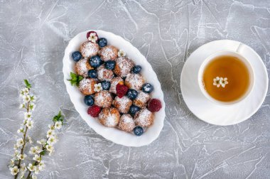Küçük kızarmış süzme peynirli çörekler oval tabakta ahududu, yabanmersini, pudra şekeri ve bir fincan çay. Kiraz çiçeği dalları gri mermer arka planda bir dekorasyon olarak kullanılır. Üst görünüm, doğal ışık, seçici odak