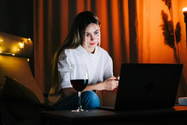 Young millennial woman having video call on laptop computer and drinking wine, use technology for communicate with friends or family.