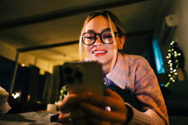 Genç gülümseyen beyaz kadın tatillerde evde, yatağın üzerinde cep telefonu kullanıyor..