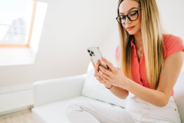 Modern evdeki büyük beyaz bir kanepede dinlenirken gözlüklü genç beyaz kadın akıllı telefon kullanıyor.. 