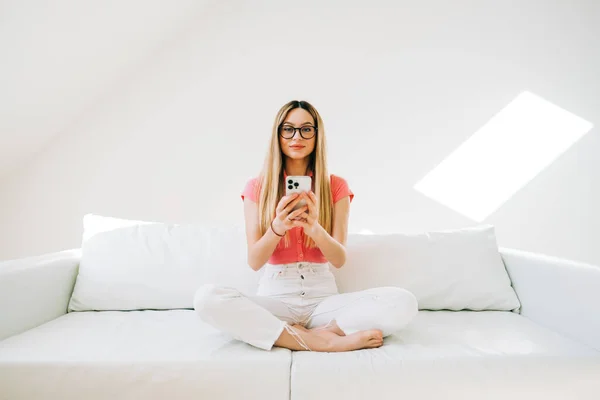 年轻的戴眼镜的高加索女人 一边拿着智能手机 一边躺在现代家里的白色大沙发上 — 图库照片