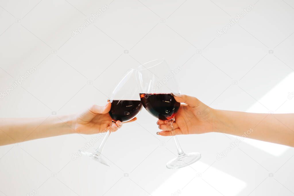 Two hands holding glasses with red whine and clinking on white wall background.