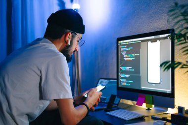 Genç adam mobil geliştirici bir bilgisayara program kodu yazar ve akıllı telefondaki uygulamayı kontrol eder, dahili ofiste programcı olarak çalışır..