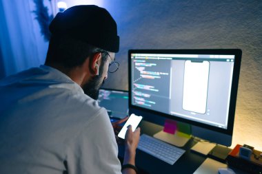 Genç adam mobil geliştirici bir bilgisayara program kodu yazar ve akıllı telefondaki uygulamayı kontrol eder, dahili ofiste programcı olarak çalışır..