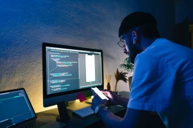 Genç adam mobil geliştirici bir bilgisayara program kodu yazar ve akıllı telefondaki uygulamayı kontrol eder, dahili ofiste programcı olarak çalışır..