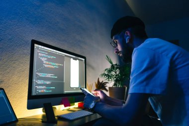 Genç adam mobil geliştirici bir bilgisayara program kodu yazar ve akıllı telefondaki uygulamayı kontrol eder, dahili ofiste programcı olarak çalışır..