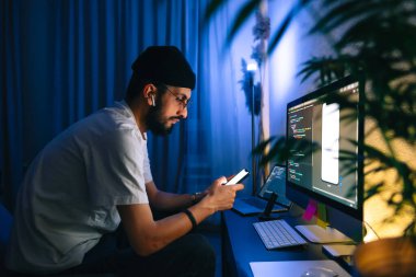 Genç adam mobil geliştirici bir bilgisayara program kodu yazar ve akıllı telefondaki uygulamayı kontrol eder, dahili ofiste programcı olarak çalışır..