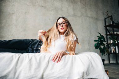 Gözlüklü, sarı saçlı, beyaz, çekici bir kadın bir yatakta oturuyor..