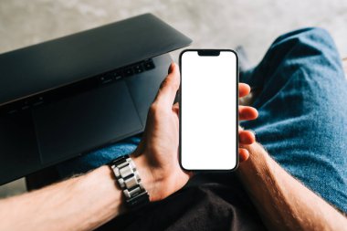 Man holding mobile phone with white screen mock up, resting on a sofa in living room at home.	