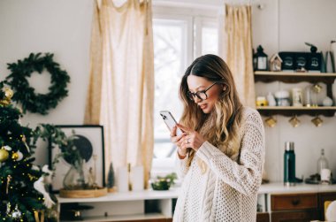 Genç beyaz kadın cep telefonu kullanıyor, evde Noel süslemeleri ile mutfakta duruyor..