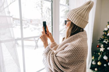 Genç beyaz kadın, sabahları pencerenin önünde dikiliyor ve kar yağdıktan sonra fotoğraf çekiyor..