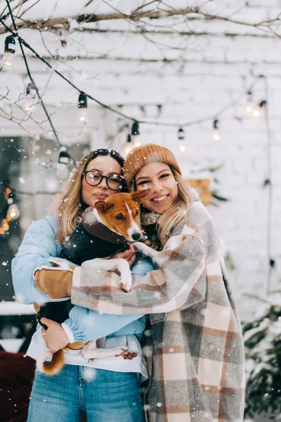 两个快乐的女性朋友站在后院 与狗在雪地里欢度冬天的肖像 — 图库照片