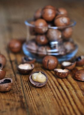 Şeffaf bir bardakta macadamia fındığı yığını ve ahşap bir yüzeyde diğerleri..