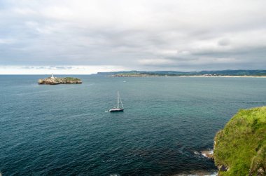 SANTANDER, SPAIN - 9 Temmuz 2021: Magdalena Yarımadası, Santander, İspanya, arka planda 19. yüzyıldan kalma deniz feneri ile küçük bir tekne yelkenlisi ve Mouro Adası 'nın görüntüsü
