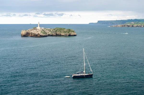 SANTANDER, SPAIN - 9 Temmuz 2021: Magdalena Yarımadası, Santander, İspanya, arka planda 19. yüzyıldan kalma deniz feneri ile küçük bir tekne yelkenlisi ve Mouro Adası 'nın görüntüsü