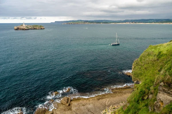 SANTANDER, SPAIN - 9 Temmuz 2021: Magdalena Yarımadası, Santander, İspanya, arka planda 19. yüzyıldan kalma deniz feneri ile küçük bir tekne yelkenlisi ve Mouro Adası 'nın görüntüsü