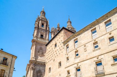 Salamanca 'da mimarlık, Castilla y Leon, İspanya. Casa de las Conchas 'ın cephesi 300' den fazla top mermisi ve arka planda bir Barok kilisesi ile dekore edilmiş geç dönem Gotik ve Plateresk tarzını karıştıran bir bina.