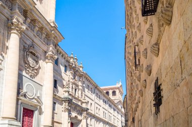 Salamanca 'da mimarlık, Castilla y Leon, İspanya. Casa de las Conchas 'ın cephesi 300' den fazla top mermisi ve arka planda bir Barok kilisesi ile dekore edilmiş geç dönem Gotik ve Plateresk tarzını karıştıran bir bina.