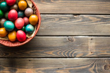 Tahta bir masanın üstünde renkli Paskalya yumurtaları olan kırmızı hasır bir sepetin üst görüntüsü.
