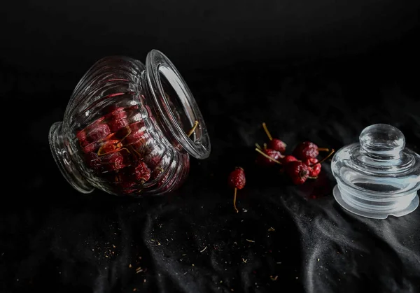 a decorative glass vase lies with an open lid filled with rose hips on a black background, close-up