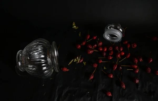a decorative glass vase lies with an open lid filled with rose hips on a black background