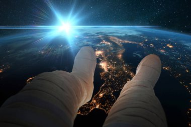 Uzaylı adam oturur ve uzaydan gelen şehirlerin gece ışıklarıyla mavi gezegen Dünya 'ya bakar. Astronot ayakları mavi günbatımında yeryüzünün arka planında. Kozmos duvar kağıdı. Güneş doğarken yörüngede dinlenen adam