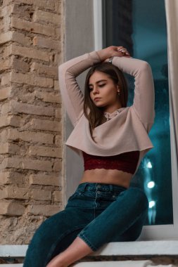 Oldukça seksi genç bir bayan model mavi moda kot pantolonlu şık pembe bir sweatshirt ile odada eski moda bir duvarın yanında pencere kenarına poz veriyor. Çekici moda kız model içeride rahatlıyor..