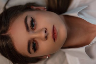Havalı, beyaz gömlekli, seksi dudakları ve doğal makyajı olan, harika gözleri olan güzel bir genç kadın. Güzel kız yatak odasındaki yatakta uzanıyor. Kadın yüzü yakın plan.
