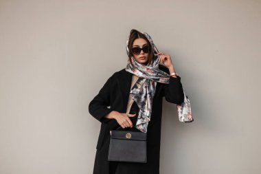 Güzel bir iş kadını modeli. Kafasında zarif bir eşarp, modaya uygun güneş gözlükleriyle, deri siyah çantalı, antika bir duvarın yanında duruyor. Seksi, lüks bir kız. Modern bayan..