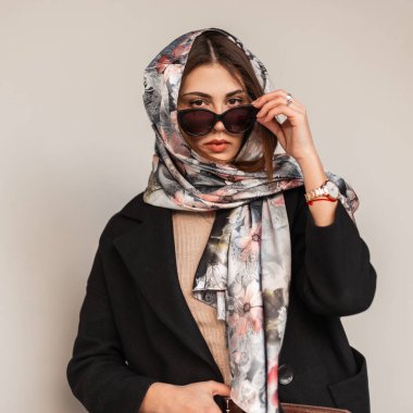Şık, ipek eşarplı, şık, siyah ceketli, şehirde poz veren şık güneş gözlüklü, zarif bir genç kadın. Modern kız model klasik beyaz duvarın yanında duruyor..