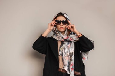 Şatafatlı, çekici, seksi dudakları olan, şık ipek eşarplı, şık siyah gözlüklü, şehirde duvarın yanında poz veren genç bir kadın. Şık şehirli kız model açık havada..