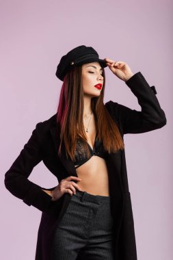 Uzun, modaya uygun ceketli, şık iç çamaşırlı, havalı kırmızı dudaklı seksi genç kadın duvarın yanındaki stüdyoda şık siyah şapkayı düzeltiyor. Modern kız model iç mekanda.