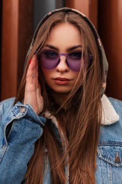 Modern metalik kırmızı duvarın yanında, havalı, mor gözlüklü, moda gençliğinin büyük boy kot ceketli havalı hippi genç kadın portresi. Avrupalı kız kentte moda kamuflaj mahallelerini düzeltiyor.