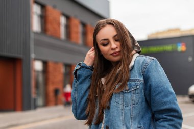 Sıradan mavi kot ceketli çekici genç bir kadın sokakta kapüşonunu düzeltiyor. Şık bir manken şehirde poz veren güzel bir kız. Kadınlar için gençlik modası kıyafetleri. Bahar stili.