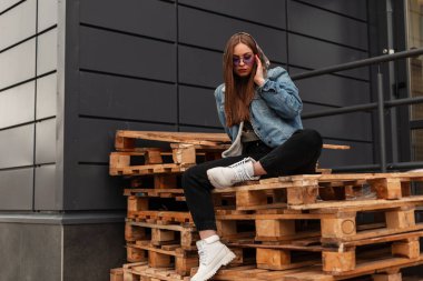 Şık kot elbiseli, şık mor gözlüklü, deri çizmeli, havalı Amerikalı genç, havalı hippi kadın şehirdeki duvarın yanındaki ahşap paletlerde dinleniyor. Çekici bir kız. Sıradan stil