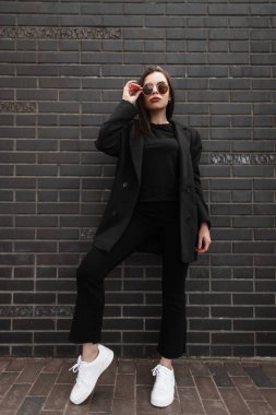 Moda tişörtlü, şık kot pantolonlu, modaya uygun siyah ceketli, şık genç bir kadın şehirde siyah tuğla duvarın yanında duruyor. Kadın vücuduna yakın çekim. Kadın kıyafetlerinin bahar koleksiyonu.