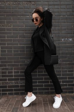 Kapalı giyim tarzı, modaya uygun uzun ceketli, kot pantolonlu, tişörtlü, sokakta tuğla binanın yanında şık, siyah çantalı bir kadın. Moda bir bahar kıyafeti. Sıradan moda..