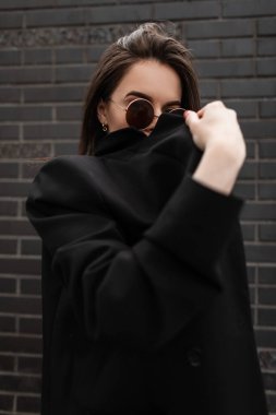 Oldukça şık, genç esmer, şık, genç, yuvarlak güneş gözlüklü bir kadın siyah, eski moda tuğla duvara karşı modaya uygun ceketle yüzünü kapatıyor. Açık havada poz veren çekici modern kız modeli. Sokak modası.
