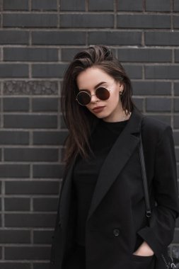 Taze portre seksi, yuvarlak güneş gözlüklü, siyah, antika ceketli, kentin tuğla duvarının yanında şık bir tişörtü olan genç bir kadın. Moda mankeni olan güzel bir kız açık havada giyiniyor..
