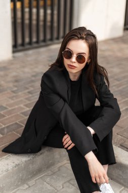 Taze portre, seksi dudakları, temiz, sağlıklı bir teni, şık bir ceketi ve şehirde yuvarlak güneş gözlüğü takan genç bir kadın. Siyah elbiseli şık kız mankeni sokaktaki antika fayanslarda dinleniyor..