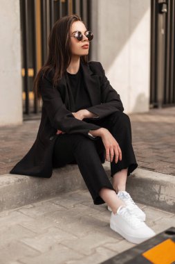 Süslü, genç, şık, siyah giyimli, spor ceketli, pantolonlu ve beyaz ayakkabılı bir kız sokakta oturuyor.