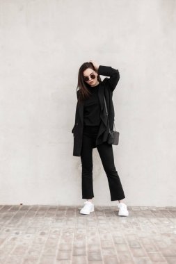 Moda güneş gözlüklü, havalı siyah gençlik kıyafetleriyle, beyaz spor ayakkabılı, şehrin klasik duvarının yanında poz veren güzel bir kadın. Çekici moda kızı. Kadınlar için gençlik kıyafetleri.