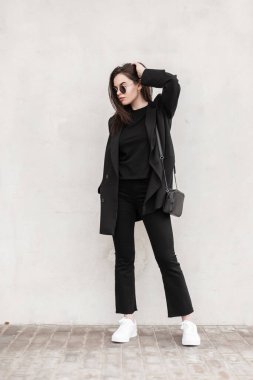 Moda mankeni, güneş gözlüklü, genç, sıradan siyah elbiseli, güzel beyaz spor ayakkabılı, sokakta klasik duvarın yanında poz veren genç bir kadın. Açık havada siyah kıyafetli Avrupalı bir şehir kızı. Sokak görünümü.