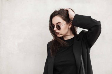 Genç gözlüklü şık genç bir kadın kentteki klasik beyaz duvarın yanında siyah elbiseli, şık güneş gözlüklü ve çantalı poz veriyor. Moda mankeni modern kız açık havada düzeliyor. Gençlik sokak stili.